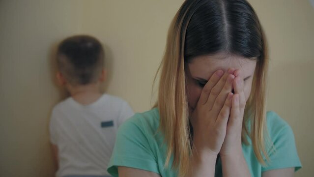 Close up: sad crying mom covers his face with hands. Resentment quarrel between mother and little son, boy sit in corner, turned away from each other. Family scandal, disagreement, soreness feeling