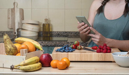 Woman looking at her cell phone in her kitchen, on a wooden table to prepare a healthy snack with fruits.
