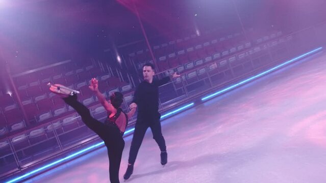 Pair of ice skaters pass by camera,man moving backwards holds by hand woman performing arabesque element, her leg and arm raised high up from behind.Concept of balance in slow motion. Camera tracking.