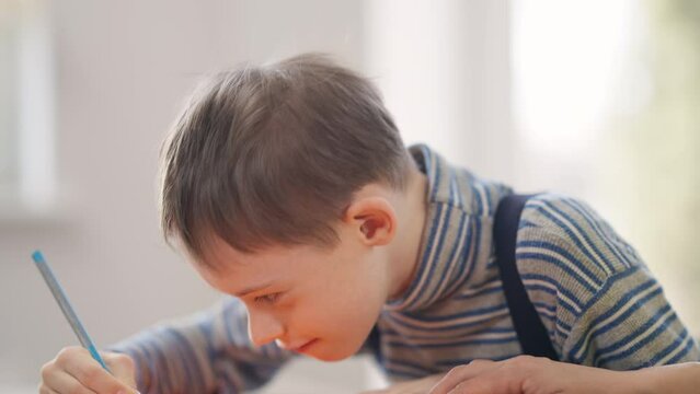 Autistic Caucasian boy painting looking away talking sitting at table in school. Portrait of child with birth anomaly genetic mutation studying art indoors. Concentration and autism concept