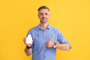 mature man with grizzled hair hold pills show thumb upon yellow background, supplement