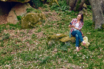 Naklejka na ściany i meble Woman sitting in forest on a stone, using mobile phone for chat or looking for gps. Traveling with backpack or exercising outdoors. Active people, hiking, healthy lifestyle and harmony concept.