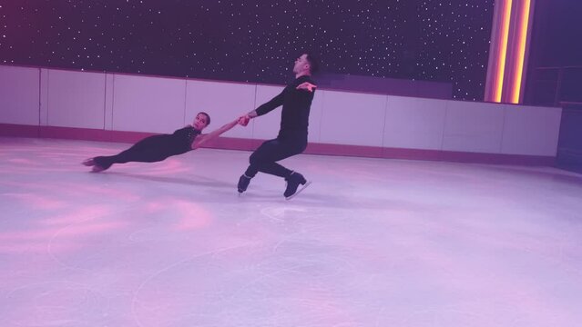 Pair of figure skaters rotate, go into death spiral,female partner hovers in air parallel to ice, man holds her hands.Figure skating element, dance support,slow motion,camera swings diagonally around.