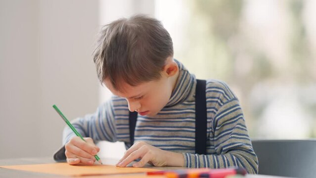 Focused autistic boy drawing with crayon on colorful paper sitting at table indoors. Portrait of concentrated Caucasian child with mental disorder genetic mutation painting in school