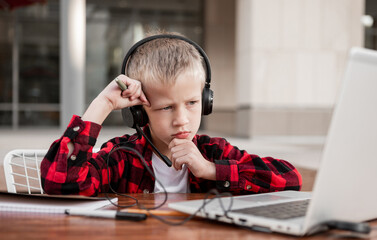 A blond boy of Slavic appearance, a schoolboy, in a checkered red shirt sits at a table in headphones.  Talking on the video link, tired and upset.  Distance learning.
