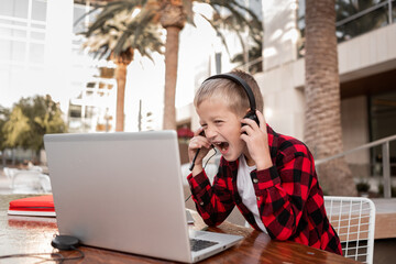 A blond schoolboy in a plaid red shirt sits at a table with headphones and a laptop.  A smiling boy...