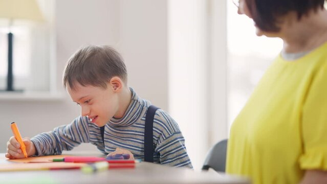 Autistic schoolboy painting talking with teacher in classroom in school. Portrait of excited curios Caucasian boy with mental illness studying indoors with blurred woman supporting child