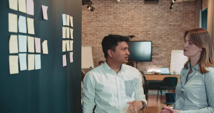 Happy multiethnic business man and woman, managers work together using sticky notes on wall at modern office slow motion