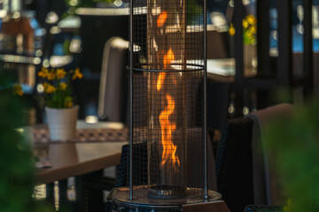 The flame of a gas burner inside glass tubes in an outdoor restaurant in the evening twilight against a blurred background of tables and yellow daffodils, selective focus