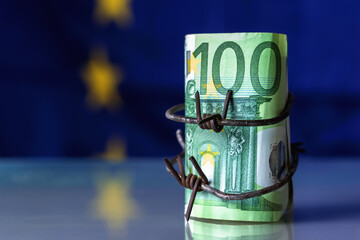 European Union currency wrapped in barbed wire against flag of EU as symbol of Economic warfare, sanctions and embargo busting. Selective focus on barbed wire. Copy space.