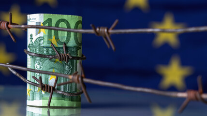 European Union currency wrapped in barbed wire against flag of EU as symbol of Economic warfare, sanctions and embargo busting. Horizontal image. Copy space.