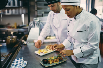 Black female chef and her colleague serving food while working at restaurant kitchen.