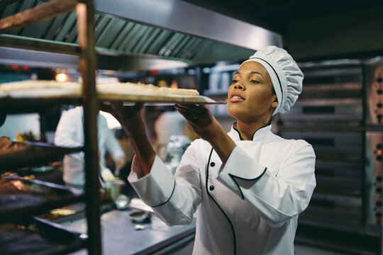 Black female chef putting tray with food on cooling rack in kitchen.
