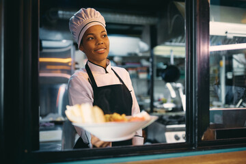 Happy African American female chef serving food through kitchen window.
