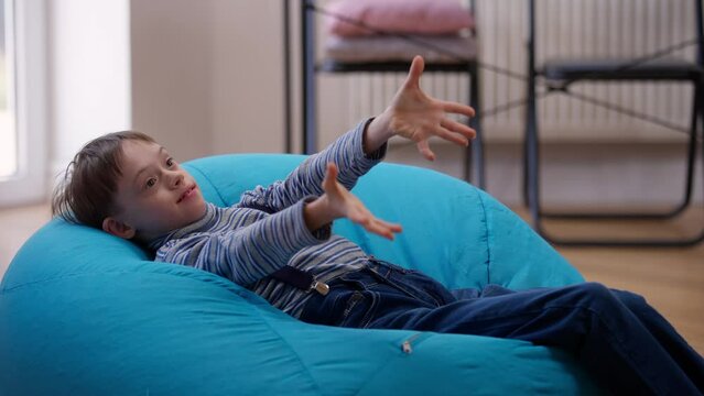 Joyful autistic boy lying on comfortable bag chair at home throwing catching teddy bear. Portrait of laughing Caucasian child with mental disorder having fun indoors. Coziness and autism concept