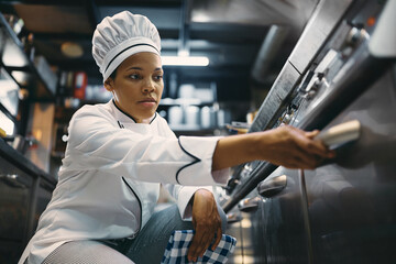 Black female professional cook using oven while preparing food at restaurant kitchen.