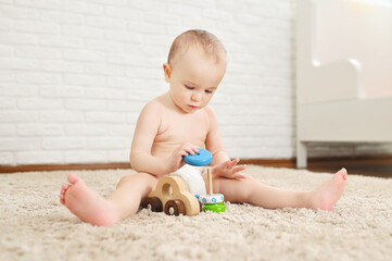 cute baby sits on a carpet in a disposable diaper plays with toys against a white brick wall.