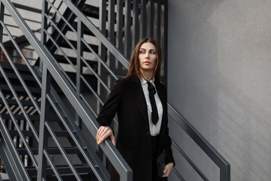Beautiful professional business woman in a fancy black suit with a shirt, tie, and coat walks down the metal stairs outside in an office building