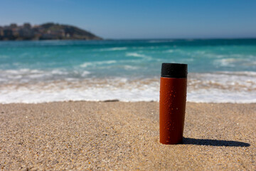 A red thermal mug on the sand by the ocean.