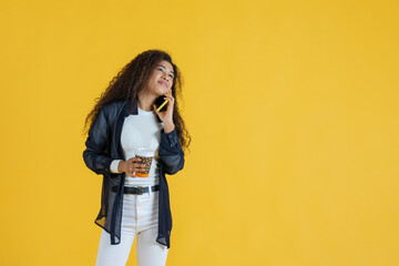 Young woman talking on phone while holding coffee cup on yellow background