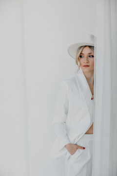 Confident young blond woman smiling, looking at camera isolated on white background. Studio portrait of successful friendly female in white suit and hat, posing over white wall.