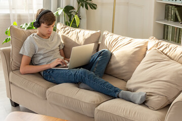 Obraz premium A teenager lying on a sofa in a room, wearing black headphones on his head, looks into a gray laptop, on his knees.