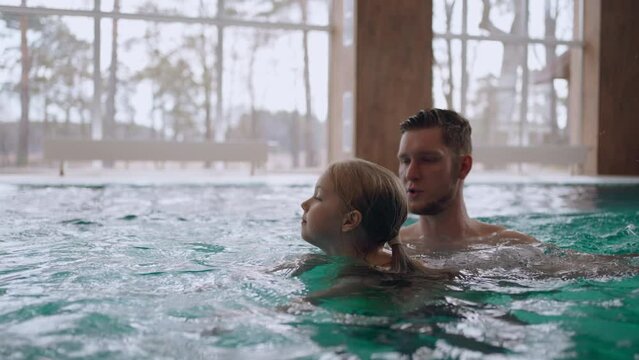 little girl is learning to swim in pool, father or swimming coach is controlling and helping her, training of swimming