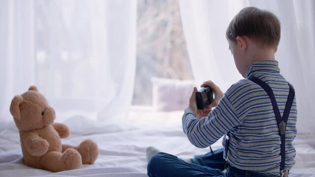 Curios autistic concentrated boy taking selfie on camera sitting on bed with teddy bear. Smiling cute Caucasian kid with mental disorder photographing himself enjoying hobby at home indoors