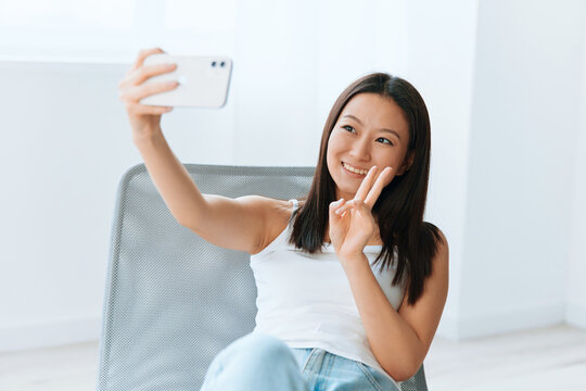 Cheerful smiling tanned lovely young Asian woman show v sign gesture doing selfie video call at home interior living room. Distance communication Social media concept. Cool offer Banner Wide angle