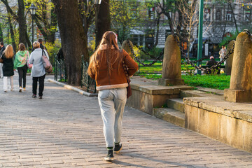 A young free girl with long hair and a pigtail walks along a spring street in the light of the evening sun with white headphones on her head behind a group of adult women with shopping bags