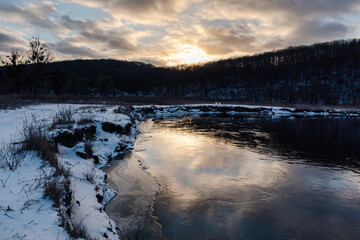 Winter view on frozen river curve with scenic sun reflection. Zmiyevsky region on Siverskyi Donets River covered in snow in Ukraine. Sunset sun in clouds above woody hill