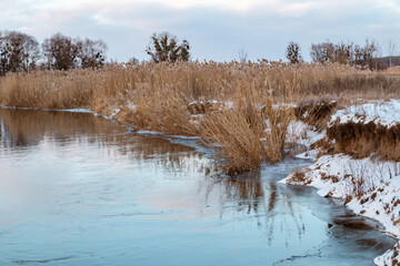 Winter view on cold river with cloudy sky with reflection in water and dry reed grass on shore. Zmiyevsky region on Siverskyi Donets River covered in snow in Ukraine