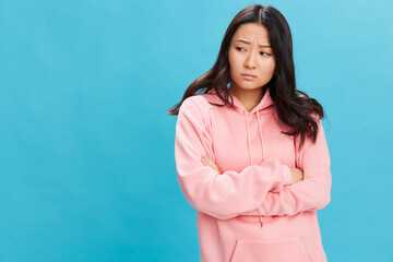 Fototapeta premium Upset sad cute Asian student young lady in pink hoodie sweatshirt folds hands posing isolated on over blue studio background. The best offer for ad. People Emotions for Everyday concept