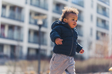 Fototapeta na wymiar Curious biracial baby boy with brown curly hair wearing warm clothes and running in his neighborhood. Outdoor shot. Sunny weather. High quality photo