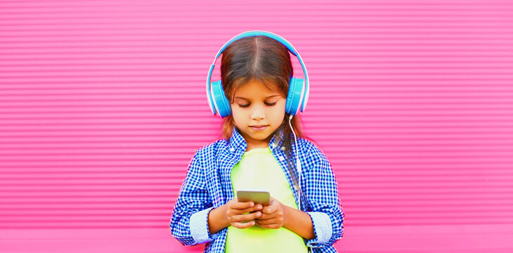 Portrait of little girl child with smartphone listening to music in wireless headphones on pink background in the city