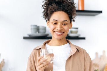 Lovely happy african american girl, in casual clothes, stands at home in the kitchen, holds a glass...