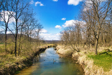 Fototapeta na wymiar Beneath a blue sky with white clouds on a Spring day, a small stream makes its way peacefully through the forests, fields and farmland of Southern Wisconsin.