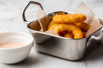 deep - fried onion rings in metal basket served with white sauce on wooden board on marble background.