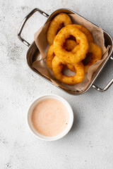deep - fried onion rings in metal basket served with white sauce on wooden board on marble background.