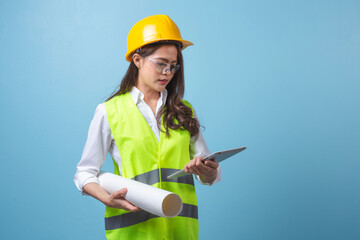 Asian woman engineer in yellow safety helmet using tablet computer standing isolated on blue background studio. Woman architect in working uniform.