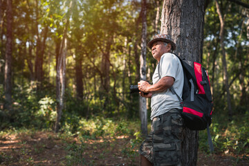 An elderly Asian man with backpack using camera while hiking in nature