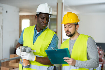 Two men construction workers in protection gear talking on indoor building construction site