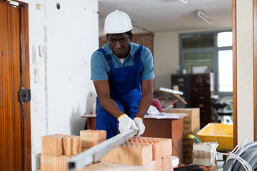 African-american man construction worker standing at brick stack and carrying metal plank in building site.