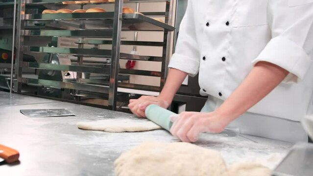 Close up of chef's hand in white cook uniforms with aprons are kneading pastry dough with roller, preparing bread, pies, and fresh bakery food, baking in oven at stainless steel kitchen of restaurant.