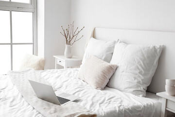 Comfortable bed with modern laptop near white wall
