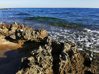 Long-hardened lava, porous, sharp coast of the Mediterranean Sea with oncoming waves against a sky.
