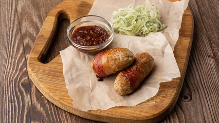 Ready-to-eat pigs sausages wrapped in bacon on wooden board. Fried savory sausages wrapped in bacon served with onion and sauce on wooden background.