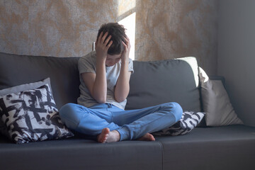 A teenage boy clutched his head with his hands, a teenage schoolboy is sitting in his room on the couch and is experiencing stress and resentment