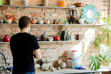 A master in a pottery shop stands against a brick wall with shelves lined with pottery.