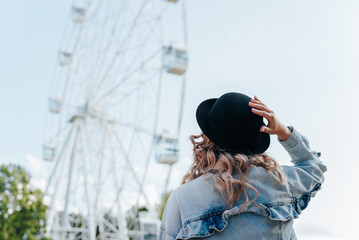 Young hipster woman in hat and denim jacket looking at ferris wheel in city amusement park on sunny...